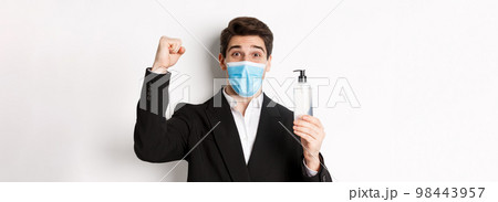 Concept of covid-19, business and social distancing. Close-up of happy man in trendy suit and medical mask, cheering and raising hand up, showing hand sanitizer, white background 98443957