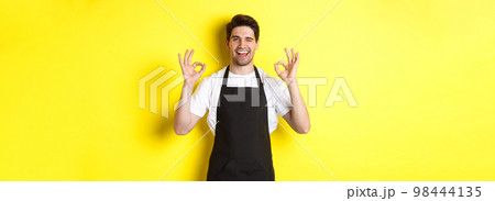 Young male barista in black apron showing OK signs and smiling, guarantee quality in coffee shop, standing over yellow background Young male barista in black apron showing OK signs and smiling, guarantee quality in coffee shop, standing over yellow background 98444135