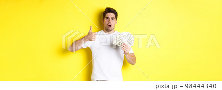 Impressed man showing thumb up, holding money credit, standing over yellow background 98444340