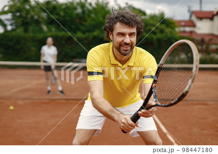 Portrait of male tennis player and female behind. Man standing on a court and playing game. Man wearing yellow t-shirt and white shorts. 98447180