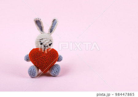 Knitted gray bunny with a crocheted red heart on a pink background. Happy Valentine's Day, Mother's Day and birthday greeting card. Knitted gray bunny with a crocheted red heart on a pink background. Happy Valentine's Day, Mother's Day and birthday greeting card. 98447865