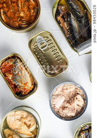 Cans with different preserve of fish and conserve seafood, opened and closed cans with Saury, mackerel, sprats, sardines, pilchard, squid, tuna,  over white stone surface top view vertical. 98450026