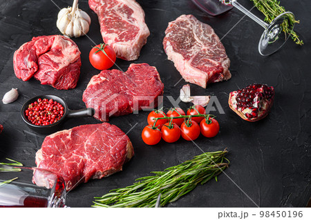 Beef steak cuts, chuck roll, rump andtop blade steak with herbs, seasoning and red wine glass on black table, side view. 98450196