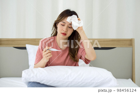 Young asian woman taking temperature with digital thermometer while sitting in bed. Covid-19 infected people stay in their rooms alone for safety, not infect people in the house. Young asian woman taking temperature with digital thermometer while sitting in bed. Covid-19 infected people stay in their rooms alone for safety, not infect people in the house. 98456124