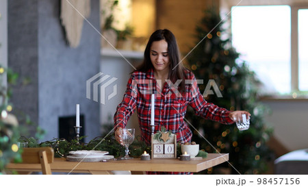 a young beautiful girl in pajamas sets the Christmas table. Puts glasses on the table. Christmas table setting. a young beautiful girl in pajamas sets the Christmas table. Puts glasses on the table. Christmas table setting. 98457156