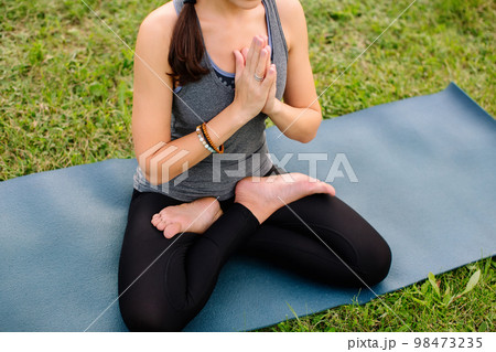 A woman instructor sitting on yoga mat in lotus position outside in city park. Namaste mudra, seated yoga asana, lotus pose, lotus posture, padmasana, cross legged sitting posture. 98473235