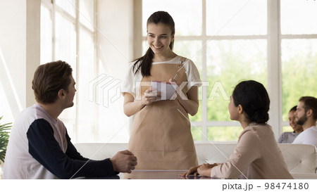 Pretty young waitress wearing uniform apron, serving diverse couple Pretty young waitress wearing uniform apron, serving diverse couple 98474180