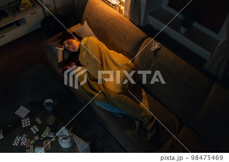 Flu and fever. Top view of illness frustrating Caucasian woman lies on sofa covered with blanket. Evening interior with electric light. Table with medicine and pills.Loneliness and depression concept Flu and fever. Top view of illness frustrating Caucasian woman lies on sofa covered with blanket. Evening interior with electric light. Table with medicine and pills.Loneliness and depression concept 98475469