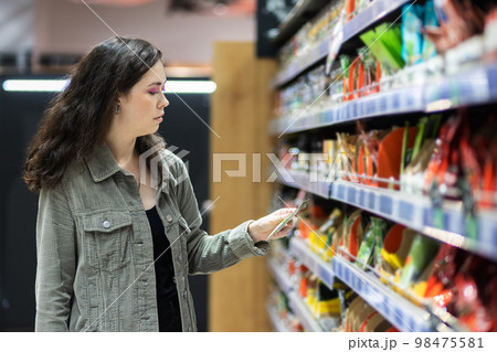 Pretty caucasian young woman choosing products on store shelf. Side view. Concept of shopping in grocery supermarket 98475581