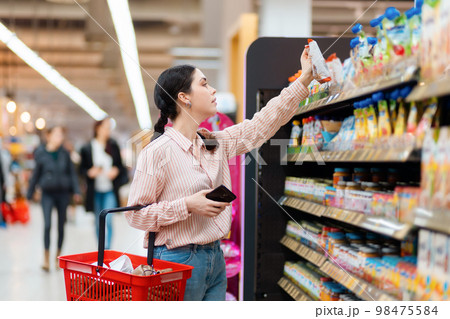 Side view of Caucasian woman choosing products in grocery store. Student reaches hand to top shelf holds cellphone and cart. Shelves with food in background. Concept of shopping and consumerism 98475584