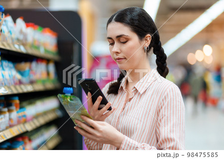 Portrait of young smiling beautiful caucasian woman scan qr code of product using smartphone. Aisle and shelves in background. Concept of shopping and consumerism 98475585