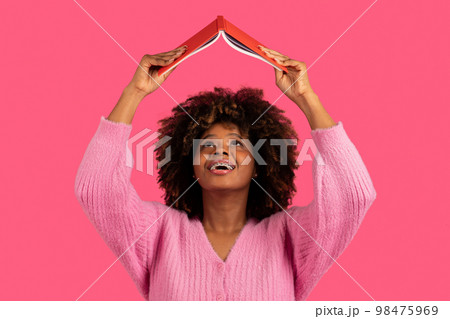 Happy funny young african american curly woman student with braces lifts book up and reads, search info 98475969