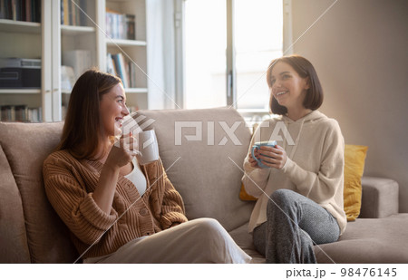 Happy european young women in casual with cups drink coffee, communication, sit on sofa in living room Happy european young women in casual with cups drink coffee, communication, sit on sofa in living room 98476145