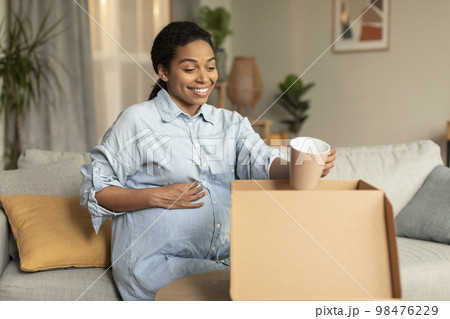 Excited pregnant black woman unpacking cardboard moving box, holding cup and smiling, sitting on couch at home 98476229