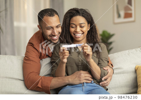 Happy african american couple holding positive pregnancy test and smiling, man hugging woman on sofa at home Happy african american couple holding positive pregnancy test and smiling, man hugging woman on sofa at home 98476230