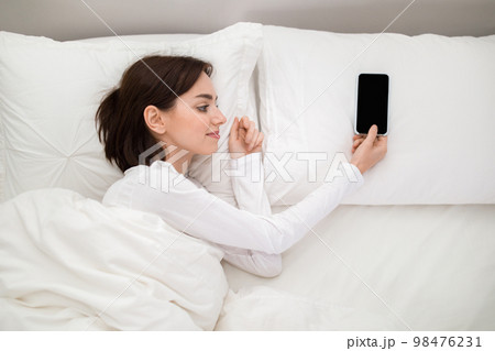 Happy young woman lying in bed with smartphone on pillow Happy young woman lying in bed with smartphone on pillow 98476231