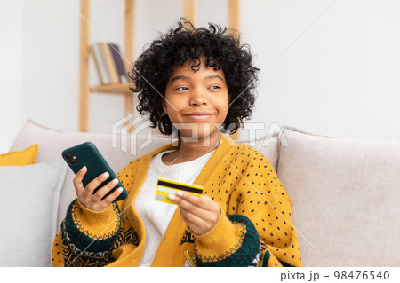 African american woman shopping online holding smartphone paying with gold credit card. Girl sitting at home buying on Internet enter credit card details. Online shopping ecommerce delivery service African american woman shopping online holding smartphone paying with gold credit card. Girl sitting at home buying on Internet enter credit card details. Online shopping ecommerce delivery service 98476540