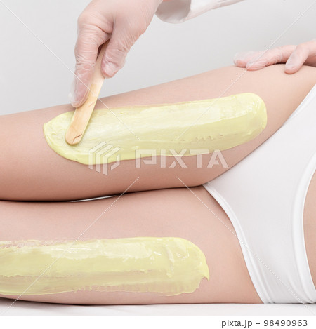 Cosmetologist in gloves applies green hot wax on slim woman legs using spatula while woman lying down on couch. Depilation with hot wax in beauty salon. Close up side view. Part of photo series. 98490963
