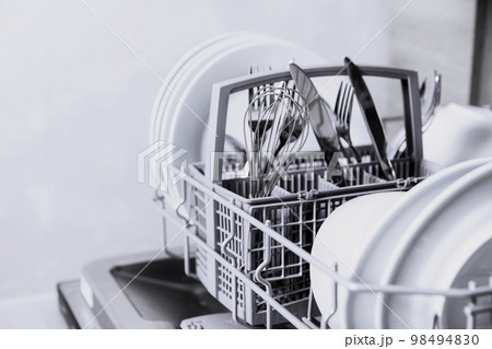 Front view of open automatic stainless steel built-in fully integrated top control dishwasher range machine with clean utensils, cutlery, glasses, dishes, plates inside in modern home kitchen 98494830
