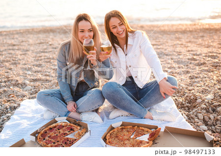 Two young beautiful girlfriends having fun with pizza and wine at beach over sea shore outdoors in sun light. Summer vacation season. Friendship. Happiness. Sisters spend time together at coast line. Two young beautiful girlfriends having fun with pizza and wine at beach over sea shore outdoors in sun light. Summer vacation season. Friendship. Happiness. Sisters spend time together at coast line. 98497133