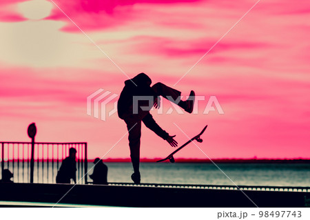 Unrecognizable teenage boy silhouette showing high jump tricks on scooter against 98497743