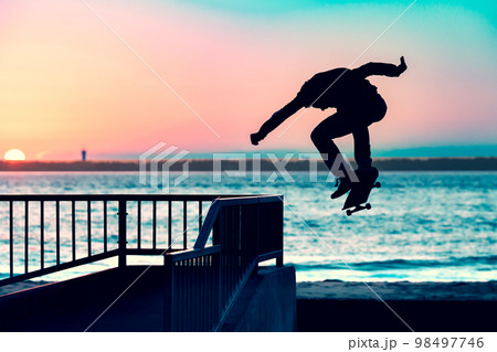 Unrecognizable teenage boy silhouette showing high jump tricks on scooter against 98497746