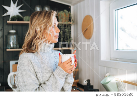 Young woman in grey sweater with cup of hot drink in hands looking at window on kitchen at house Young woman in grey sweater with cup of hot drink in hands looking at window on kitchen at house 98501630