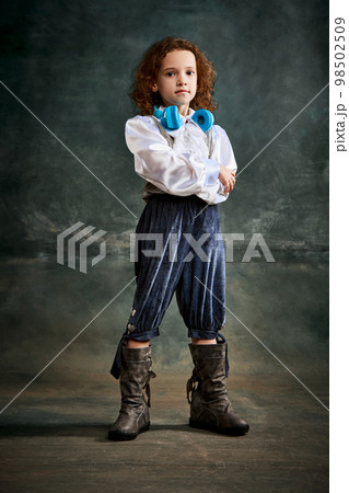Portrait of little charming girl with curly hair in image of little prince posing over on dark green background. Concept of historical remake, comparison of eras, retro, vintage, emotions Portrait of little charming girl with curly hair in image of little prince posing over on dark green background. Concept of historical remake, comparison of eras, retro, vintage, emotions 98502509
