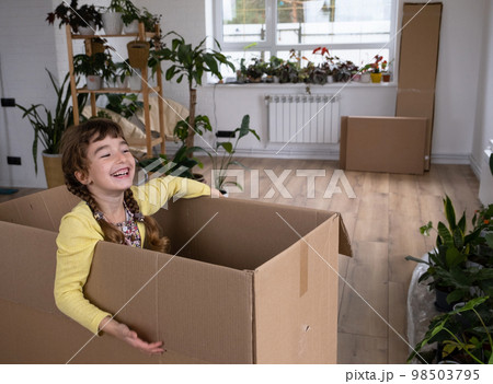 The girl hides in a big box and looks out of it funny in a room inside the home. Moving to a new house, unpacking things, renting an apartment, mortgage, housing issue The girl hides in a big box and looks out of it funny in a room inside the home. Moving to a new house, unpacking things, renting an apartment, mortgage, housing issue 98503795