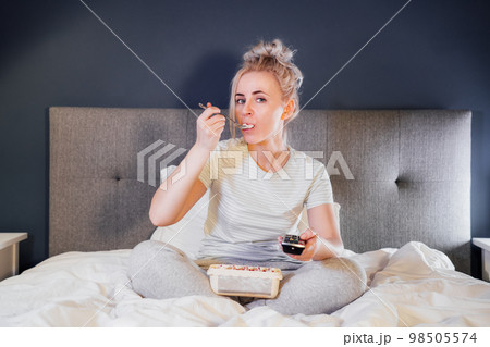 Bored, frustated woman eating ice cream and using the remote control to change the channel while watching television in bed at night. Overeating, Jamming of negative emotions. Selective focus. Bored, frustated woman eating ice cream and using the remote control to change the channel while watching television in bed at night. Overeating, Jamming of negative emotions. Selective focus. 98505574