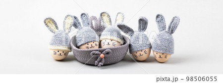 Five Easter eggs with funny faces and gray crocheted hats with bunny ears in a decorative basket on a gray background. Happy Easter concept. Banner 98506055