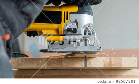 Close-up of a man cutting a wooden plank with an electric jigsaw in a workshop. Close-up of a man cutting a wooden plank with an electric jigsaw in a workshop. 98507581