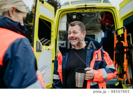Rescuers having break in front of ambulance car, talking and drinking coffee. 98507665