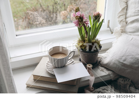 Feminine spring composition. Cup of coffee, books and blank greeting card mockup near window. Vintage Easter styled photo. Moody floral composition. Potted pink hyacinth flowers. Linen cushion. Feminine spring composition. Cup of coffee, books and blank greeting card mockup near window. Vintage Easter styled photo. Moody floral composition. Potted pink hyacinth flowers. Linen cushion. 98509404