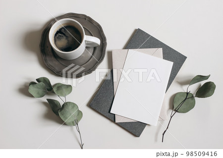Moody breakfast still life, stationery. Blank greeting card, invitation mockup, dry eucalyptus tree branches and grey diary. Cup of coffee on silver plate, white table background. Flat lay, top view. Moody breakfast still life, stationery. Blank greeting card, invitation mockup, dry eucalyptus tree branches and grey diary. Cup of coffee on silver plate, white table background. Flat lay, top view. 98509416