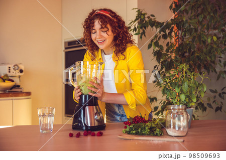 Curly-haired girl looking involved while making smoothie in the kitchen Curly-haired girl looking involved while making smoothie in the kitchen 98509693