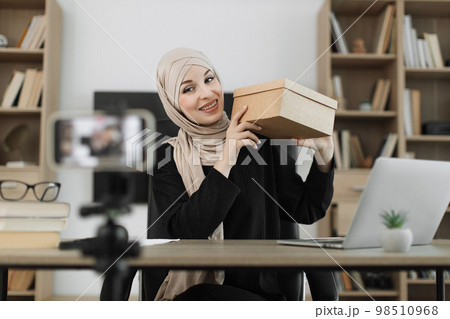 Pretty muslim woman in casual wear and hijab recording video on camera while unpacking gift boxes. 98510968