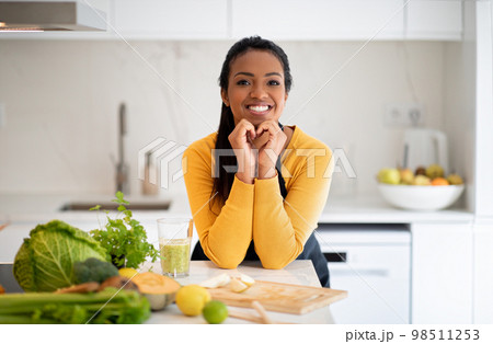 Happy young african american lady make heart sign with hands at table with organic vegetables, fresh smoothie Happy young african american lady make heart sign with hands at table with organic vegetables, fresh smoothie 98511253