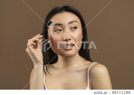 Beautiful young japanese lady brushing brows and applying eyebrow mascara, posing over brown studio background 98511286