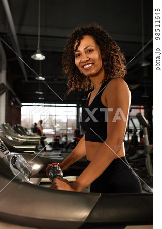Fit smiling beautiful african american woman running on treadmill in gym 98511643
