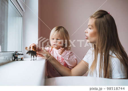 Little girl toddler with her mother playing with animal toys on window sill in children's room at home 98518744