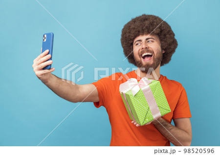 Portrait of happy joyful man with Afro hairstyle wearing orange T-shirt having video call with present box, boasting his gift to followers. Indoor studio shot isolated on blue background. 98520596