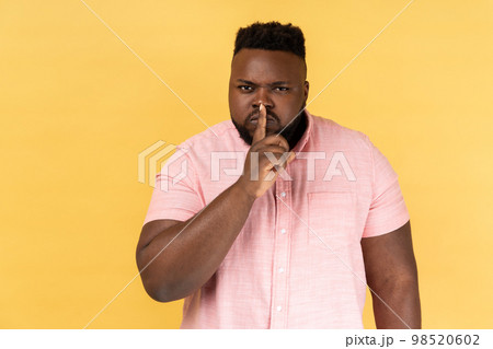Shh, make silence please. Portrait of serious strict man wearing pink shirt standing showing quiet gesture, saying hush, keep secret. Indoor studio shot isolated on yellow background. 98520602