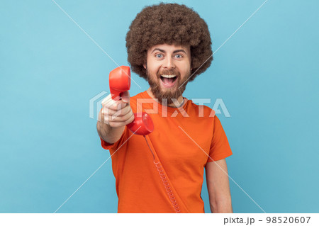 Portrait of man with Afro hairstyle wearing orange T-shirt holding and showing retro phone handset to camera, asking to answer phone. Indoor studio shot isolated on blue background. Portrait of man with Afro hairstyle wearing orange T-shirt holding and showing retro phone handset to camera, asking to answer phone. Indoor studio shot isolated on blue background. 98520607