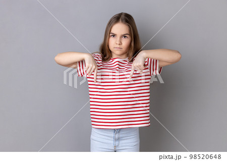 Here and right now. Portrait of serious bossy dark haired little girl wearing striped T-shirt standing and pointing index fingers down. Indoor studio shot isolated on gray background. Here and right now. Portrait of serious bossy dark haired little girl wearing striped T-shirt standing and pointing index fingers down. Indoor studio shot isolated on gray background. 98520648