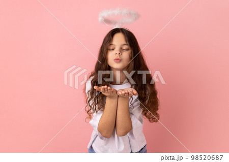 Portrait of little girl wearing white T-shirt with halo on head sending air kiss on camera, flirting, valentines day, keeps eyes closed. Indoor studio shot isolated on pink background. 98520687