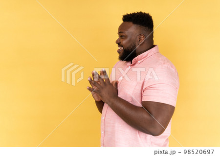 Side view portrait of cunning bearded man wearing pink shirt clasping hands and planning evil tricky prank or scheming, cheating with sly smile. Indoor studio shot isolated on yellow background. Side view portrait of cunning bearded man wearing pink shirt clasping hands and planning evil tricky prank or scheming, cheating with sly smile. Indoor studio shot isolated on yellow background. 98520697