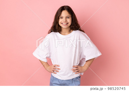 Portrait of delighted smiling little girl wearing white T-shirt standing hands on hips, looking at camera with happy expression. Indoor studio shot isolated on pink background. 98520745