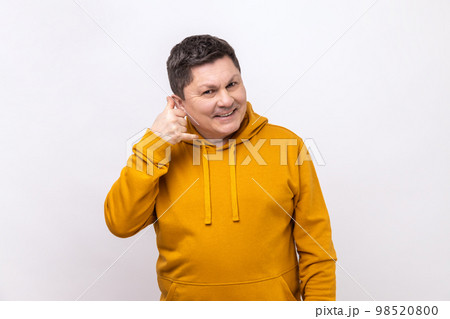Portrait of handsome cheerful man holding telephone hand gesture near ear, imitating communication on phone, wearing urban style hoodie. Indoor studio shot isolated on white background. 98520800