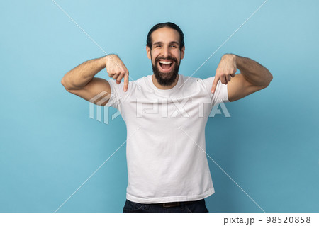 Look at ads below. Portrait of man with beard wearing white T-shirt smiling and pointing down, showing place for idea presentation, commercial text. Indoor studio shot isolated on blue background. 98520858
